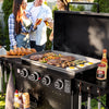 A black outdoor Royal Gourmet gas grill with a large flat top griddle is shown in a backyard setting with people blurred in the background. The grill is loaded with steak, mushrooms, peppers, eggs, and toasted bread, with condiment bottles hanging and placed on the sides.