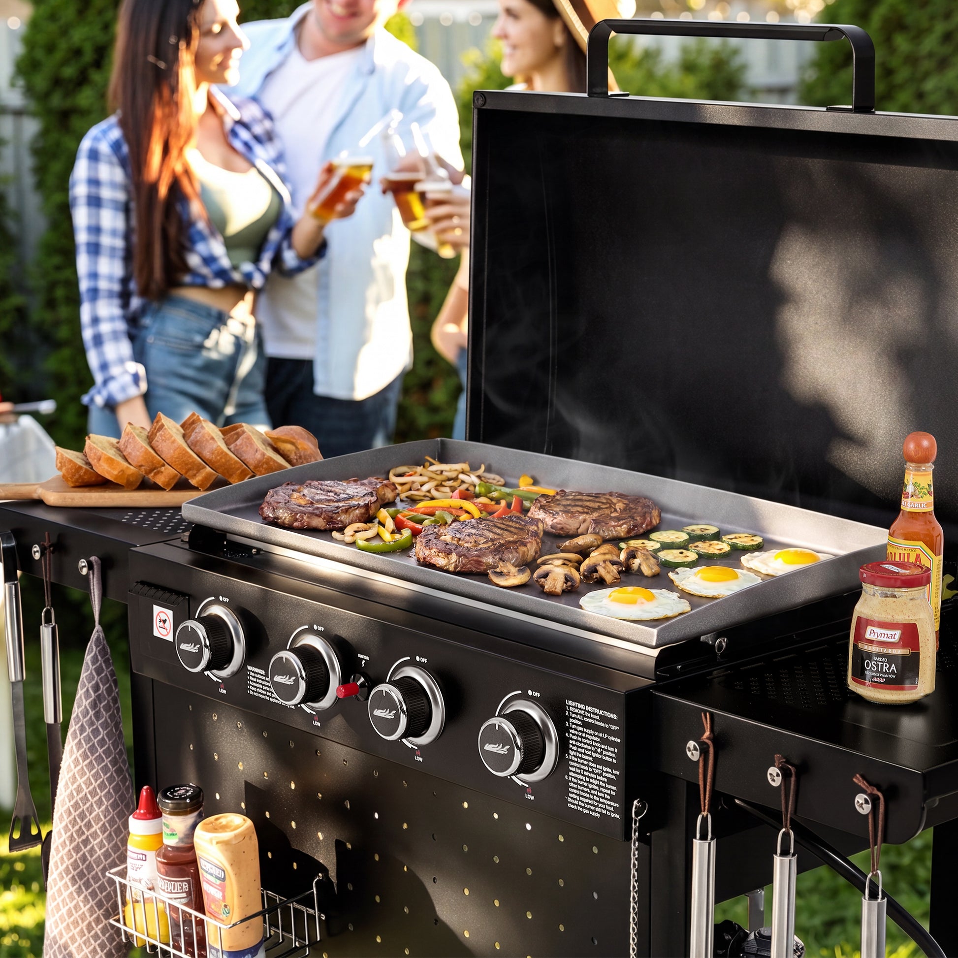 A black outdoor Royal Gourmet gas grill with a large flat top griddle is shown in a backyard setting with people blurred in the background. The grill is loaded with steak, mushrooms, peppers, eggs, and toasted bread, with condiment bottles hanging and placed on the sides.