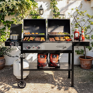 A large Royal Gourmet dual-grill outdoor charcoal grill set up on a patio, with food cooking on both grates, a propane tank underneath, and bags of charcoal on the lower shelf. Condiment bottles sit on the side table, and plants and a white wall are visible in the background.