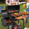 A black outdoor Royal Gourmet pellet grill with an open lid and side table, shown cooking ribs, sausages, corn, and kebabs on a grassy lawn. Two people are partially visible behind the grill, one holding an orange drink.