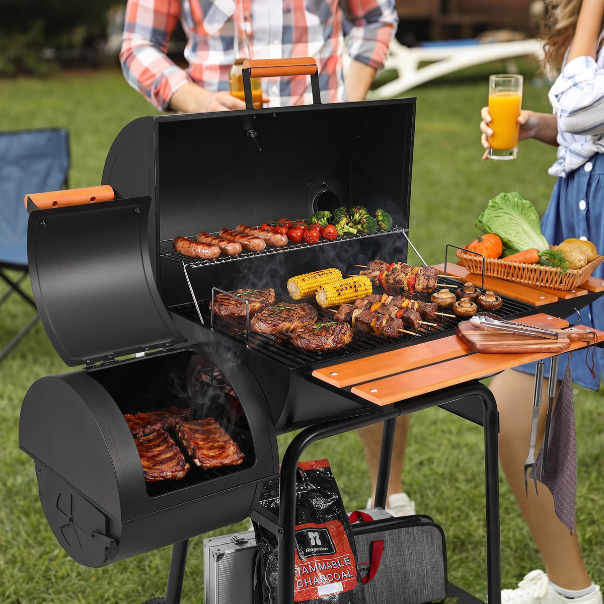 A black outdoor Royal Gourmet pellet grill with an open lid and side table, shown cooking ribs, sausages, corn, and kebabs on a grassy lawn. Two people are partially visible behind the grill, one holding an orange drink.