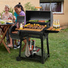 A black Royal Gourmet  charcoal grill with a lid is set up on a grassy backyard patio, loaded with food on the grates and serving shelves. Two women are seated at a dining table to the left, and a house exterior is visible in the background.