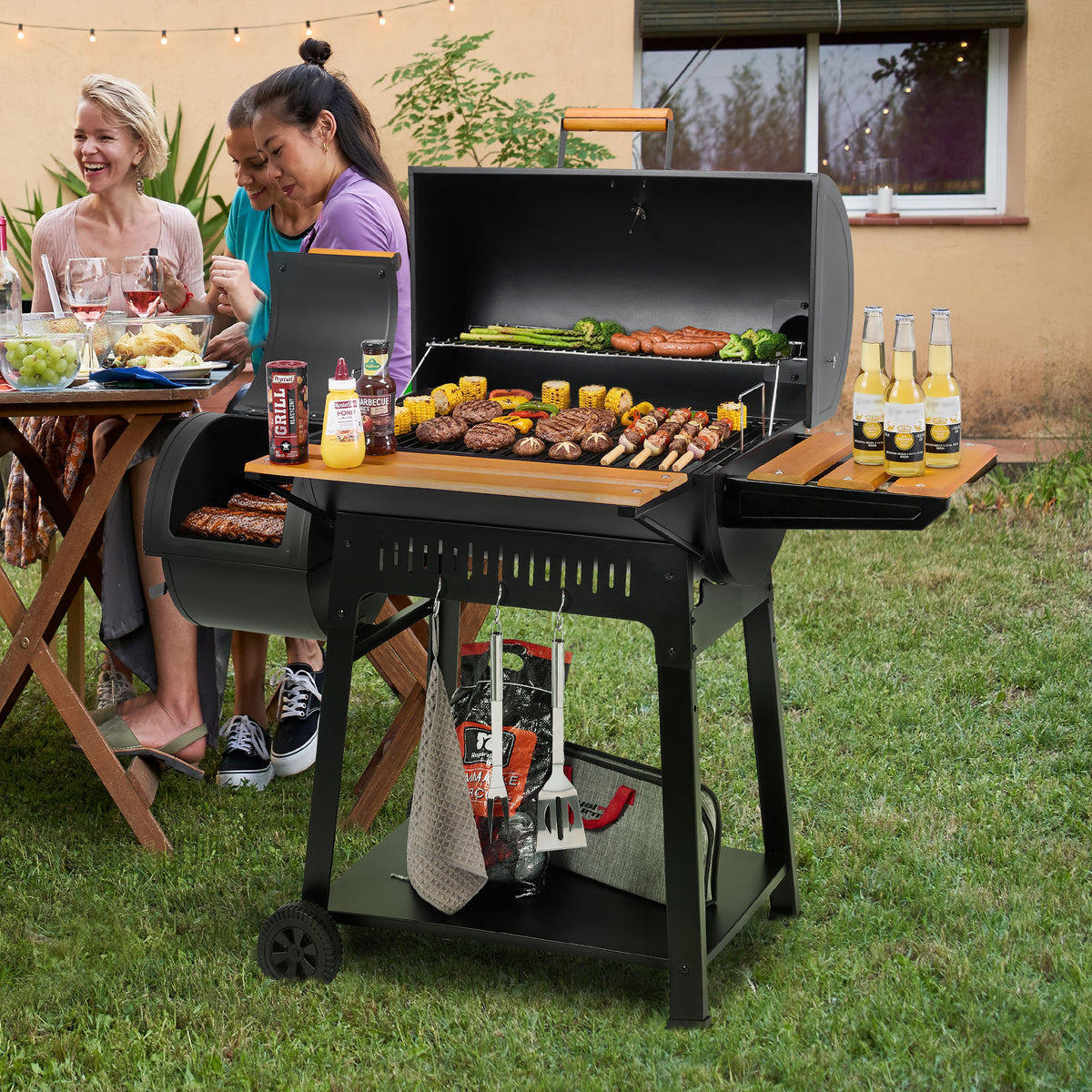 A black Royal Gourmet  charcoal grill with a lid is set up on a grassy backyard patio, loaded with food on the grates and serving shelves. Two women are seated at a dining table to the left, and a house exterior is visible in the background.