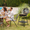 A black outdoor Royal Gourmet charcoal grill with an open lid is set on a grassy lawn next to a couple sitting at a table by a swimming pool. The scene is set in a real environment with people present, making the subject_type 'Lifestyle'.