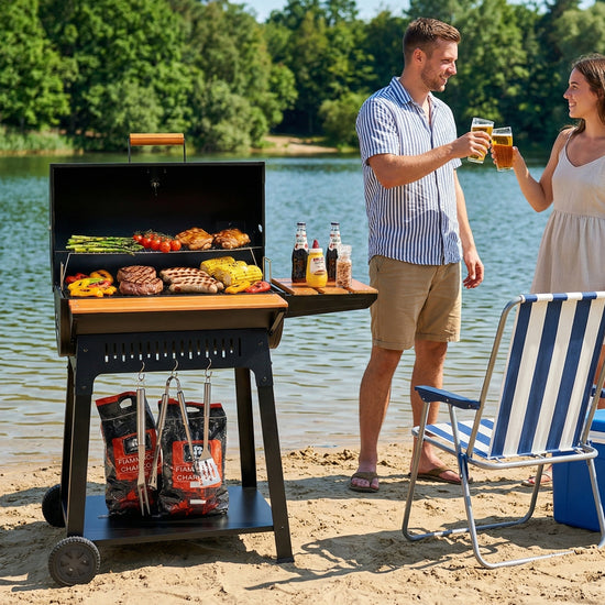 A black Royal Gourmet outdoor charcoal grill with a side table is set on a sandy lakeside patio, cooking grilled meats and vegetables. A man and a woman are standing behind the grill, clinking glasses.