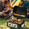A portable black Royal Gourmet gas grill with a multi-zone griddle is shown open on grass, cooking sausages, pineapple, mushrooms, asparagus, and toast. A person sitting behind plays an acoustic guitar, with a yellow and blue tent and camping gear in the background.