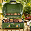 An olive green portable Royal Gourmet gas grill with food cooking inside, set on a wooden table with a blurred garden and outdoor dining background.