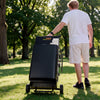 A man in a white t-shirt and beige shorts pushes a black wheeled cart filled with books or binders through a sunny, grassy park with trees.