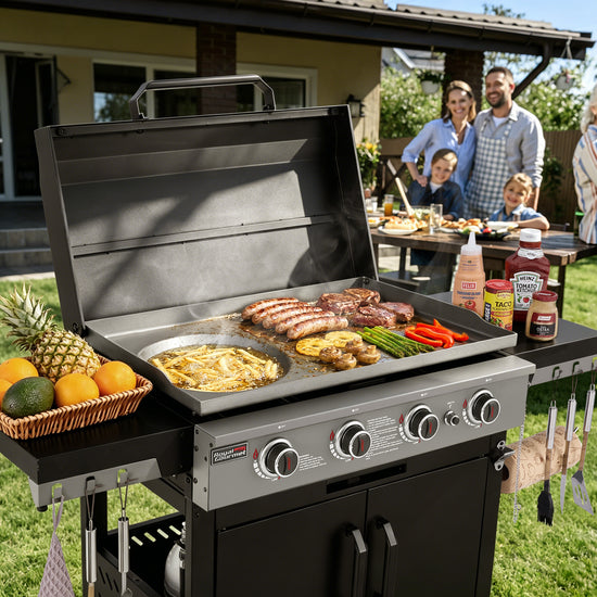 A black and stainless steel outdoor Royal Gourmet  gas grill is shown open in a backyard setting, cooking sausages, steaks, burgers, asparagus, and vegetables. A basket of fruit sits on the side table, and a family is visible dining in the background.