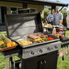 A black and stainless steel outdoor Royal Gourmet  gas grill is shown open in a backyard setting, cooking sausages, steaks, burgers, asparagus, and vegetables. A basket of fruit sits on the side table, and a family is visible dining in the background.