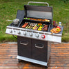A stainless steel and black Royal Gourmet gas grill with six control knobs stands on a wooden deck outdoors, cooking sausages, mushrooms, peppers, and skewers. A small pot sits on the left burner, and condiments and a cutting board with peppers are on the right side.