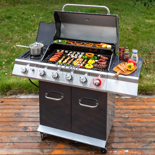 A stainless steel and black Royal Gourmet gas grill with six control knobs stands on a wooden deck outdoors, cooking sausages, mushrooms, peppers, and skewers. A small pot sits on the left burner, and condiments and a cutting board with peppers are on the right side.