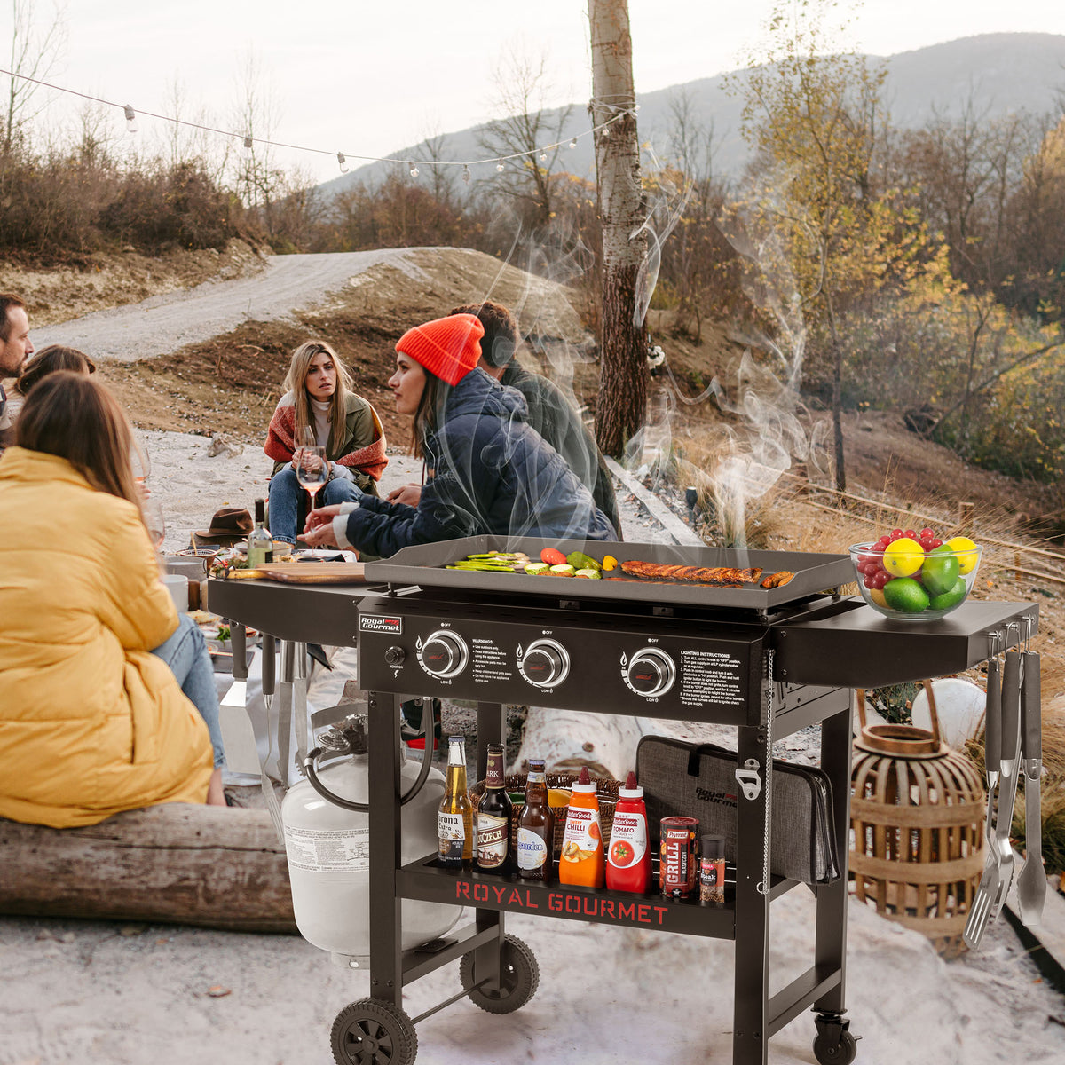 A Royal Gourmet outdoor gas grill with a flat top griddle is set up in a campsite environment with people sitting nearby and mountains in the background.