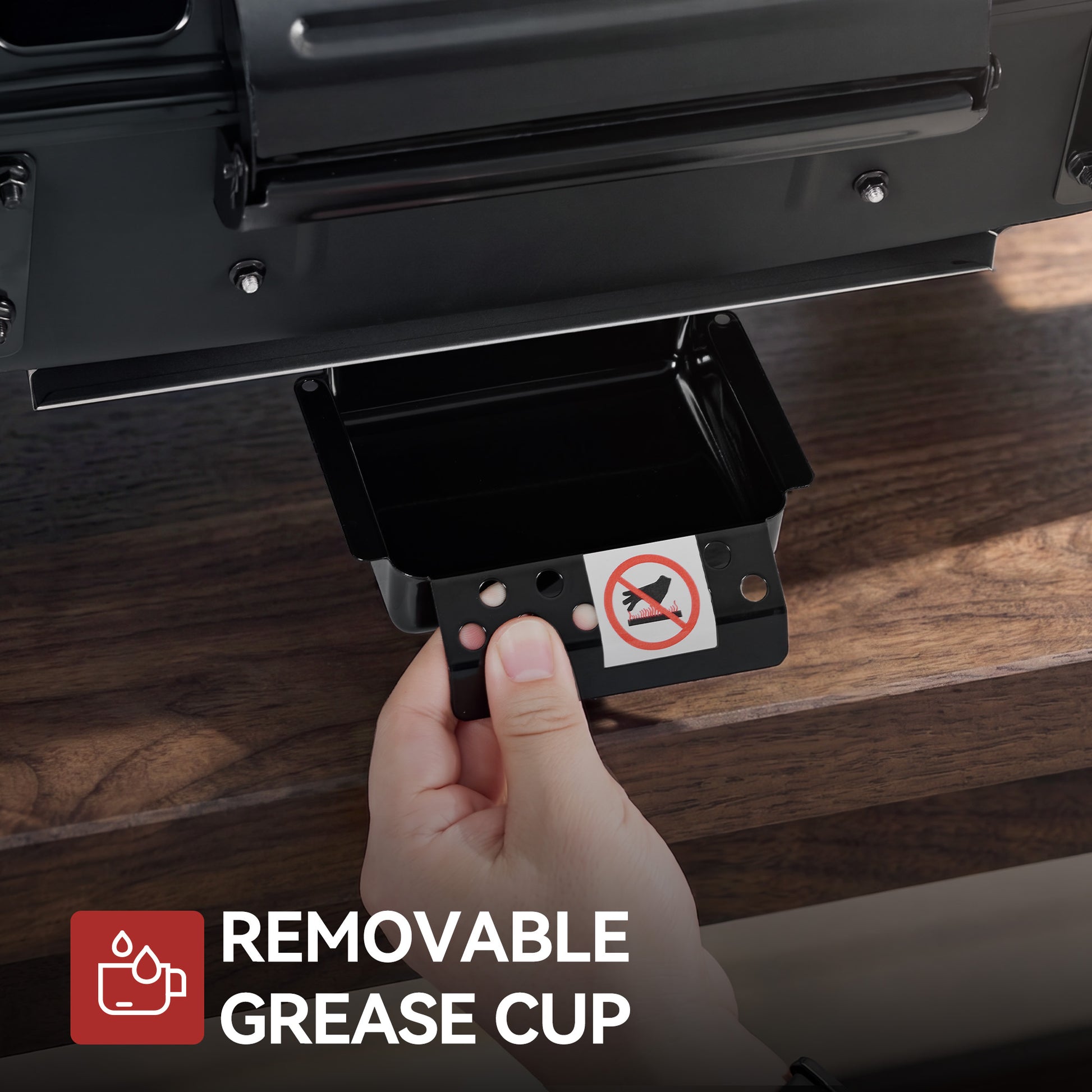 A hand holds a small Royal Gourmet black removable grease cup beneath an open compartment of a dark metal appliance, with a wooden surface in the background and a text overlay reading 'REMOVABLE GREASE CUP'.