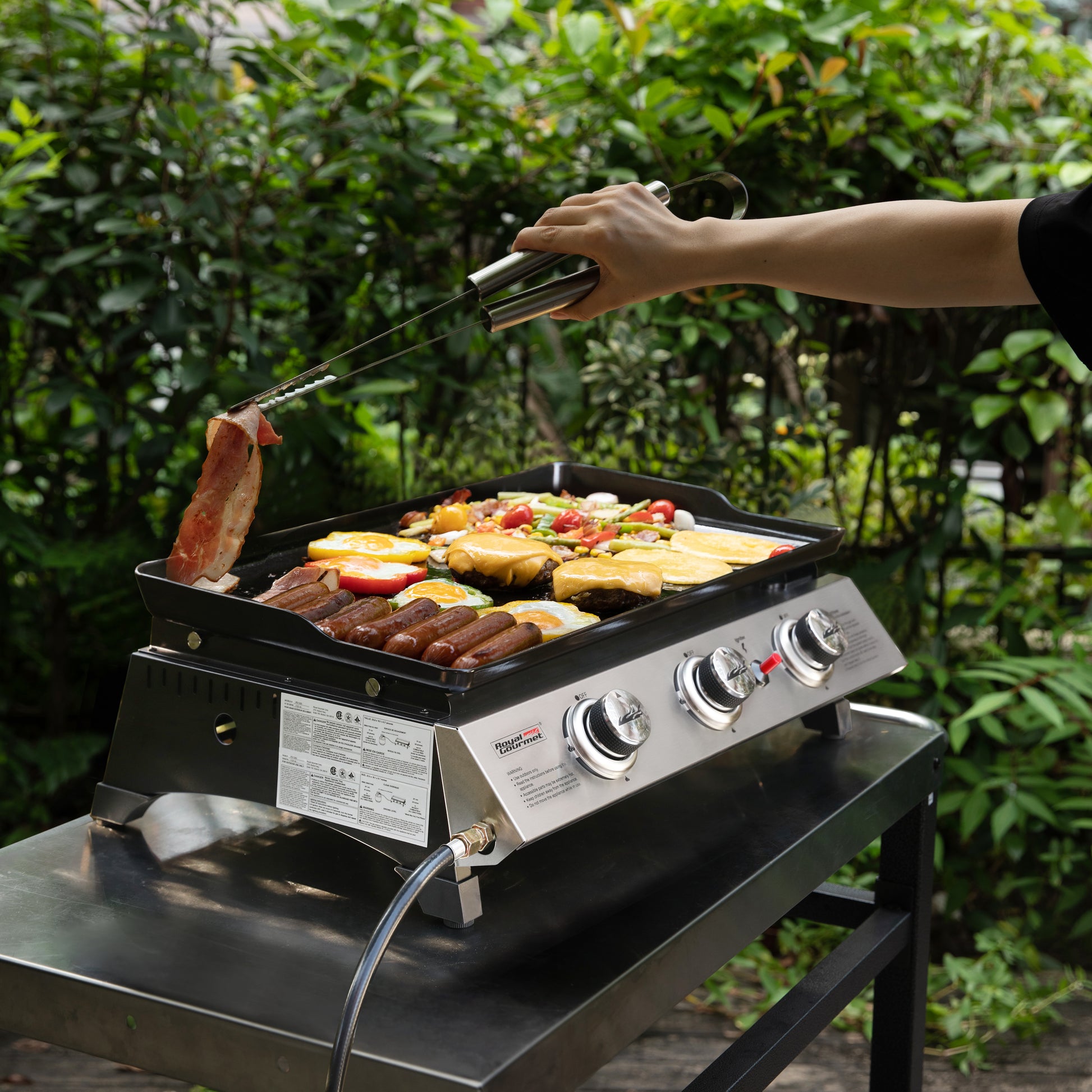 Person grilling vegetables and sausages on a Royal Gourmet portable grill outdoors.