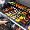 A stainless steel Royal Gourmet gas grill filled with food, including skewered vegetables, mushrooms, bell peppers, ribs, and sausages, with a person's gloved hands adjusting the lid. A metal pot is visible in the background.