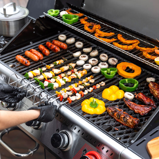 A stainless steel Royal Gourmet gas grill filled with food, including skewered vegetables, mushrooms, bell peppers, ribs, and sausages, with a person's gloved hands adjusting the lid. A metal pot is visible in the background.