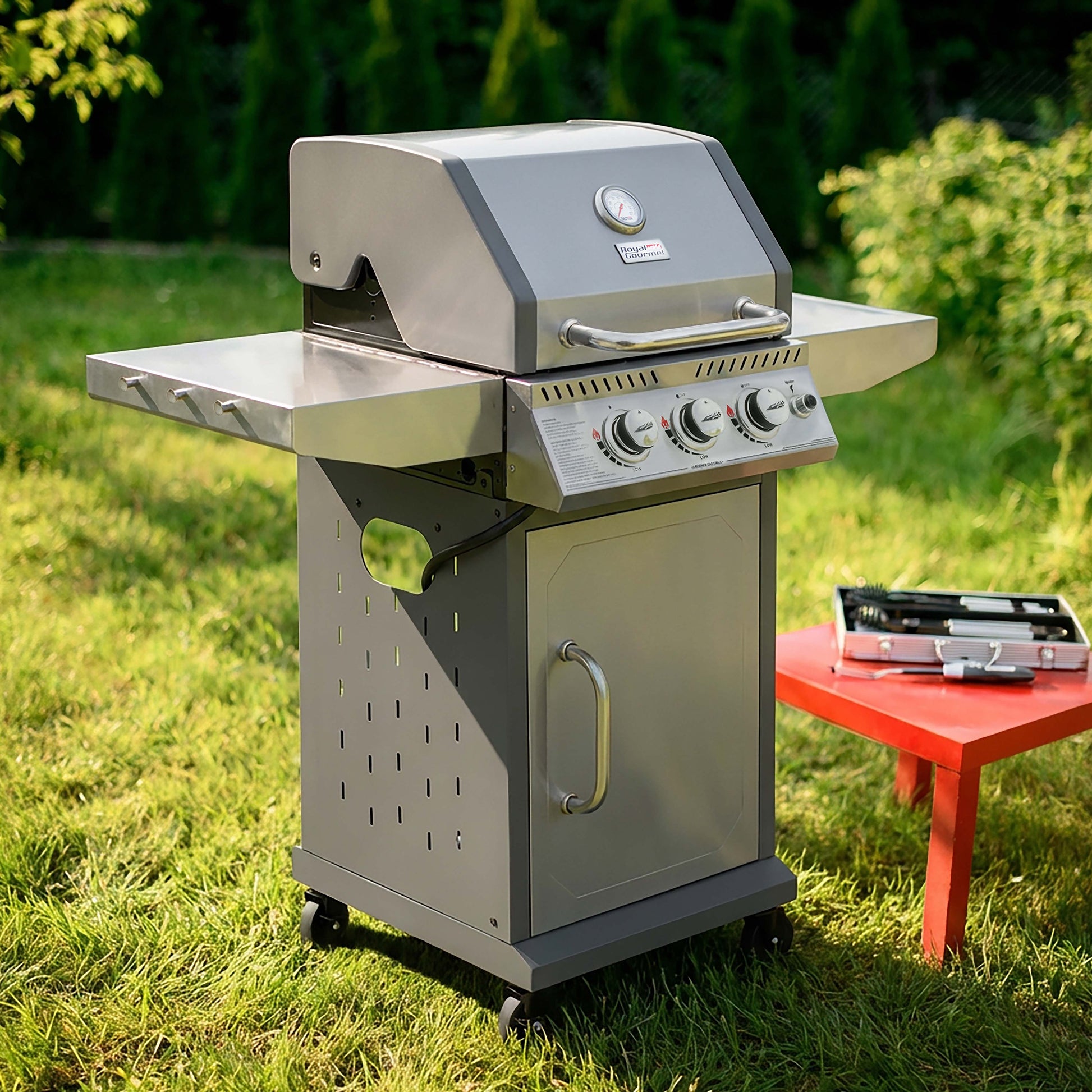 A stainless steel outdoor Royal Gourmet gas grill with side tables and a lower storage cabinet, shown on a grassy lawn next to a small red table holding a tool case.