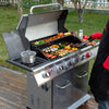 A stainless steel 5-burn Royal Gourmet  outdoor gas grill is shown on a wooden deck with food cooking inside, a person in a black apron and red shirt holding tongs at the right edge, and grass/trees in the background.