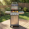 A stainless steel Royal Gourmet gas grill with side tables and a lower storage cabinet, shown outdoors on a wooden deck with food cooking inside. The grill features a brushed metal finish, four control knobs, and a handle on top, set against a background of green grass and a wooden fence.
