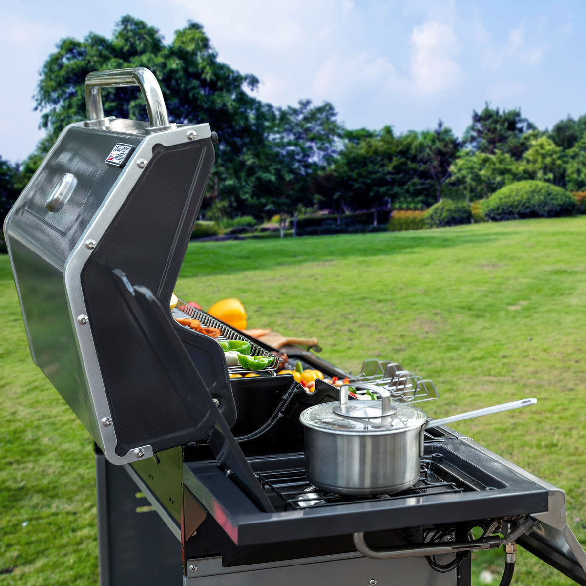 A stainless steel outdoor Royal Gourmet gas grill with its lid open, set on a grassy lawn with trees and sky in the background. A pot sits on the cooking surface, and cooking utensils and vegetables are visible inside the grill.