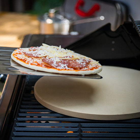 Uncooked pizza being placed on a grill with a Royal Gourmet pizza stone.