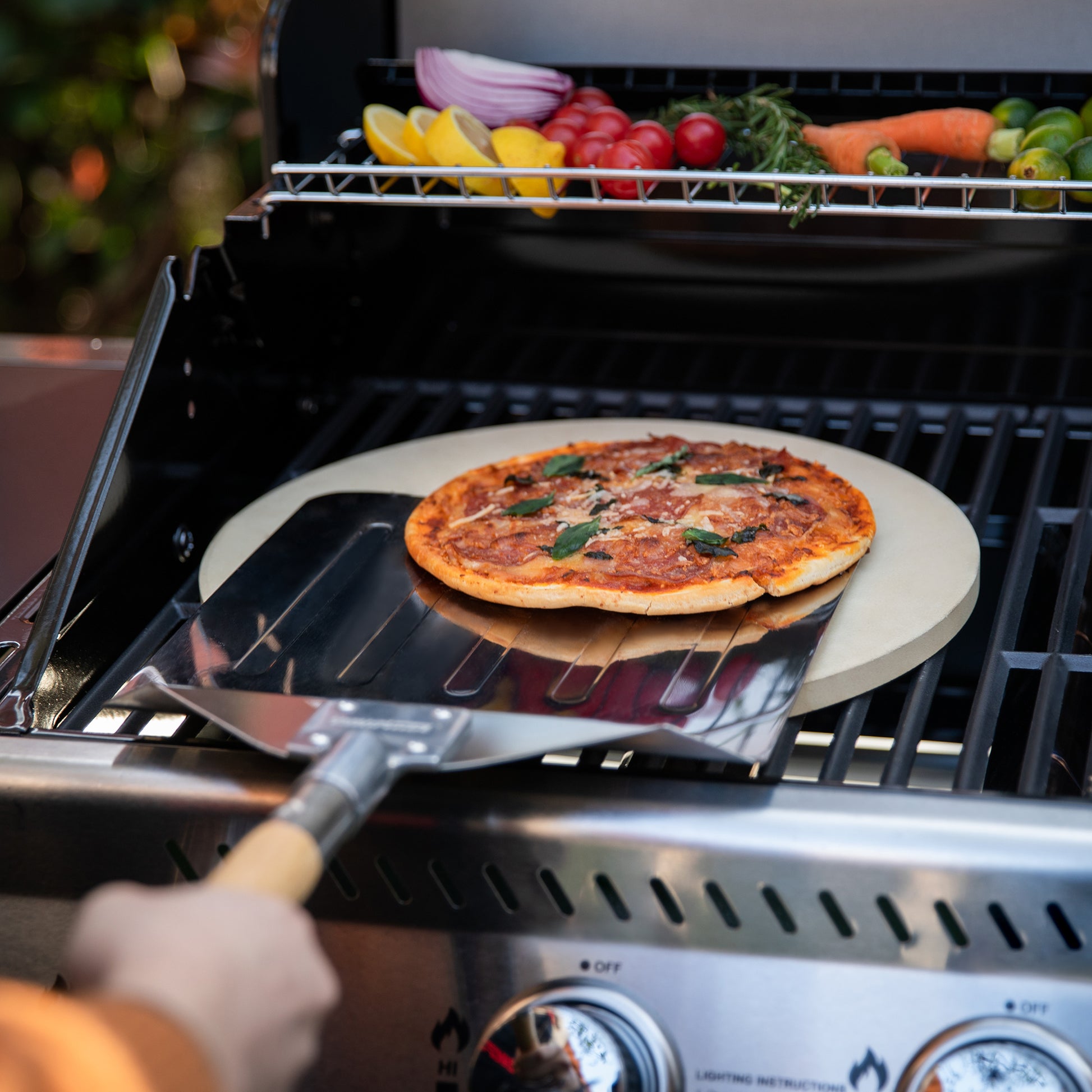 Person using a Royal Gourmet pizza peel to place a pizza on a grill with vegetables.